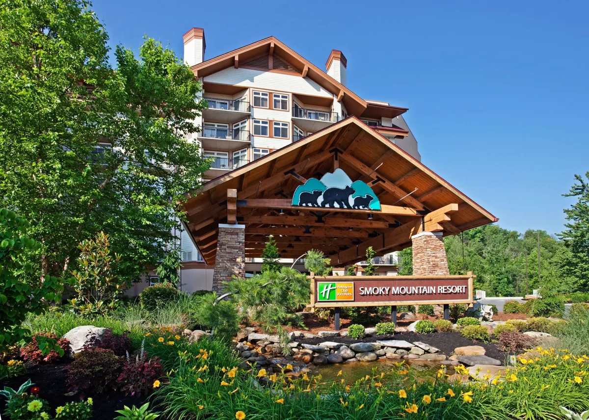 The main entrance of the Smoky Mountain Resort, featuring a large wooden canopy, stone accents, colorful flower beds, and clear resort signage. Guests are seen checking in, framed by a lush and inviting landscape, hinting at the start of an adventure-filled vacation.