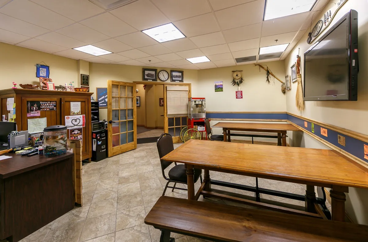 This image shows a welcoming communal dining area with wooden tables, a reception desk in the background, a TV on the wall, and decorative items that enhance the inviting atmosphere. It suggests a space where guests might gather for meals, meetings, or socializing.