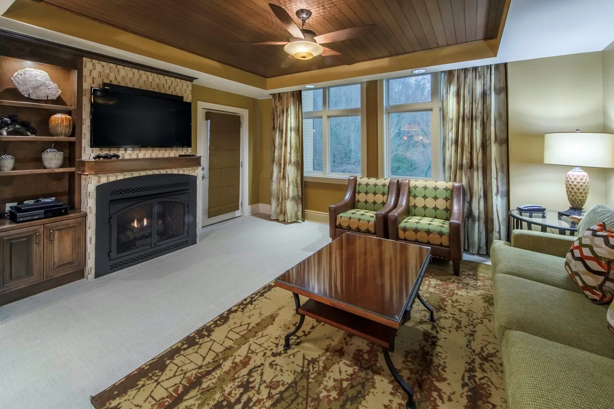 The image features a cozy living area with a glowing fireplace as the centerpiece, surrounded by plush seating and a coffee table. Large windows offer glimpses of the lush Smoky Mountains beyond, while the inviting space invites guests to gather, relax, and unwind after a day of adventure.