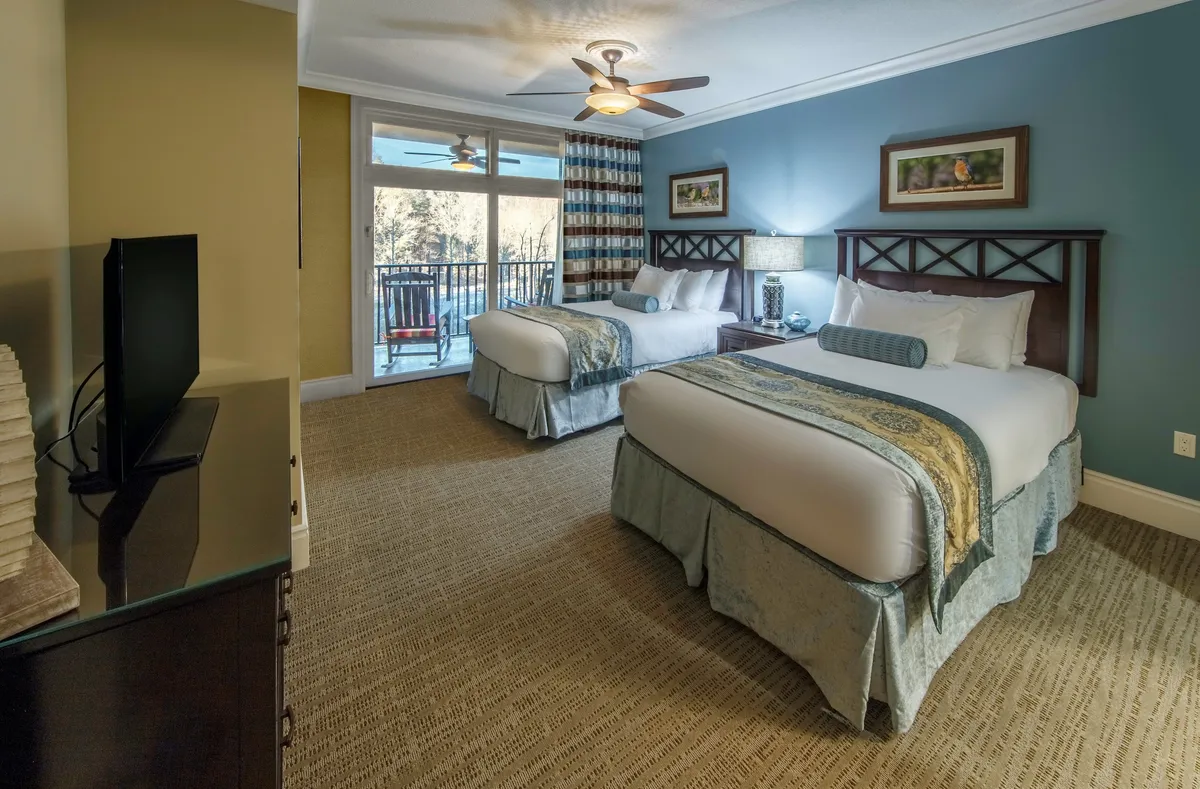 The second bedroom offers two comfortable double beds adorned with decorative bed skirts. A large window provides access to a balcony, filling the room with natural light. The setting is completed by a television and tasteful wall art, ensuring a welcoming space for children or friends.