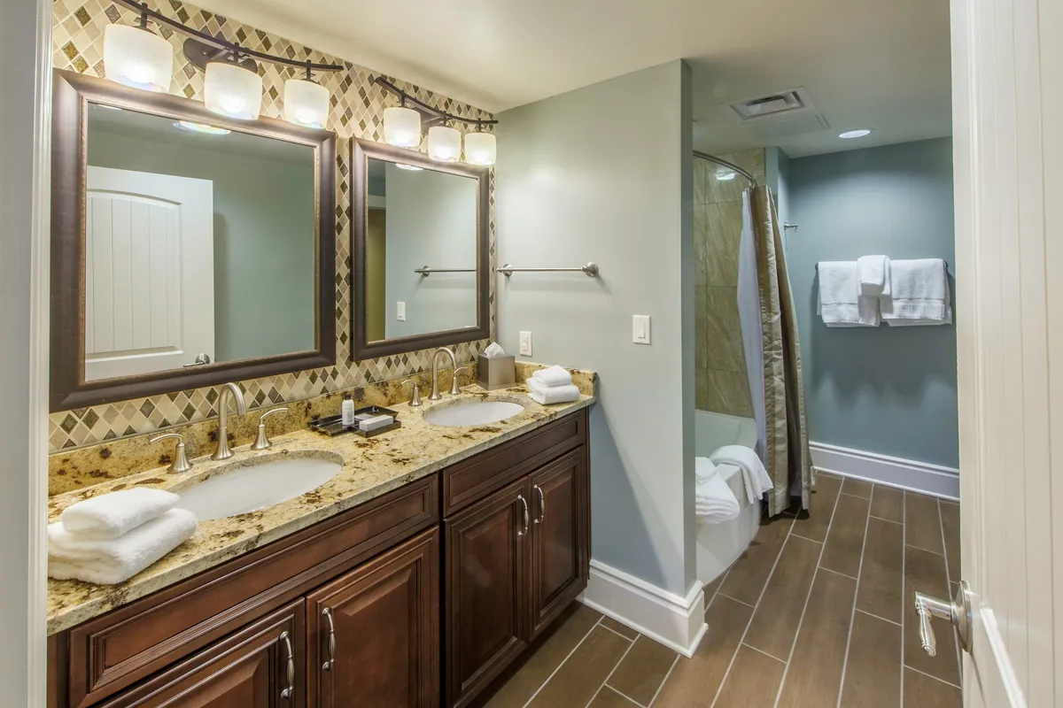 The spa-inspired bathroom features elegant double vanities with a granite countertop, large mirrors, and a beautifully tiled shower area. The space exudes a serene, luxurious feel, perfect for pampering and self-care.